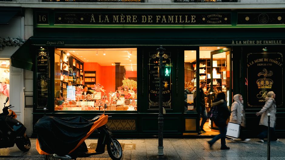 A cozy view of a Paris chocolate shop warmly lit, enticing pedestrians and shoppers.