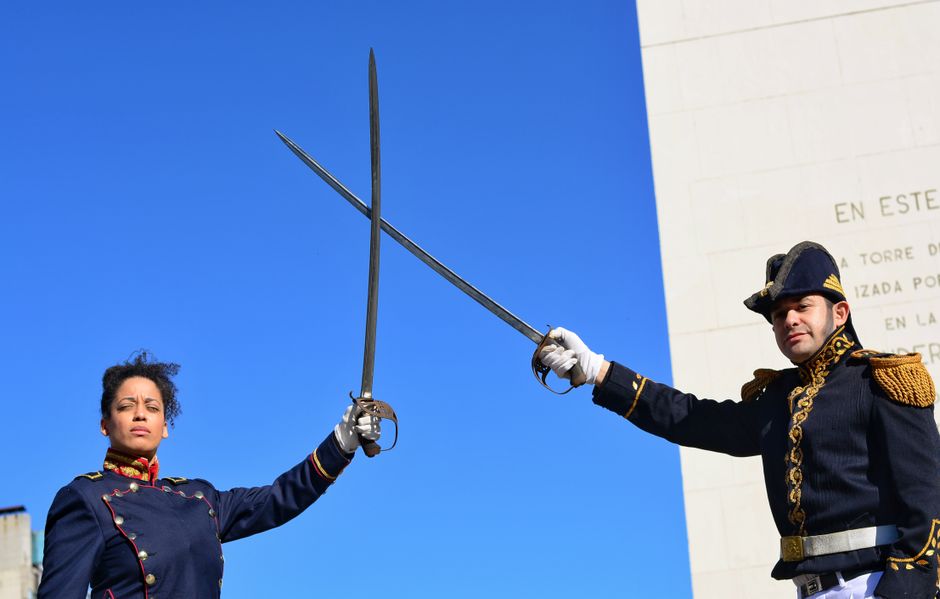 Two actors in period uniforms reenacting a duel with swords held high under a clear blue sky.