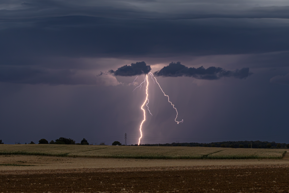 A powerful lightning bolt illuminates the cloudy evening sky over a rural field in France.