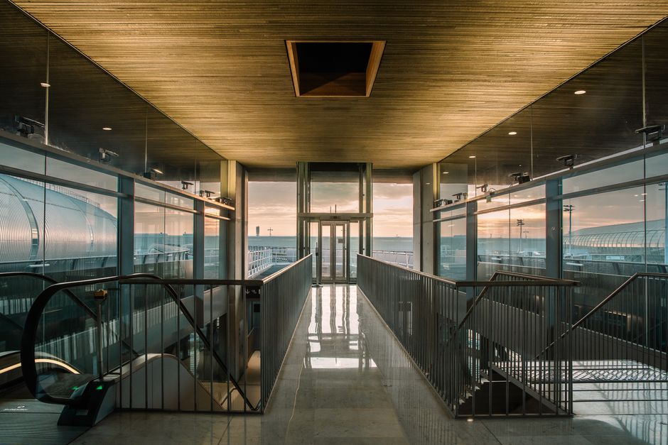 A modern airport terminal corridor with glass walls, overlooking the outdoor area at sunrise.