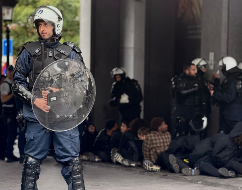 Riot police guard a protest scene in Brussels with detained individuals seated nearby.