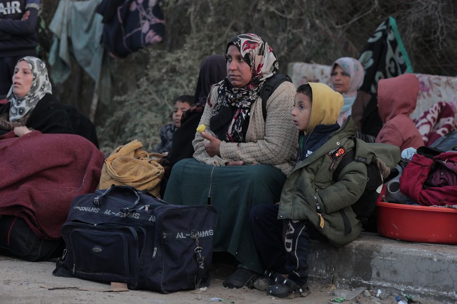 Adults and children sit outdoors in a refugee camp, surrounded by belongings.