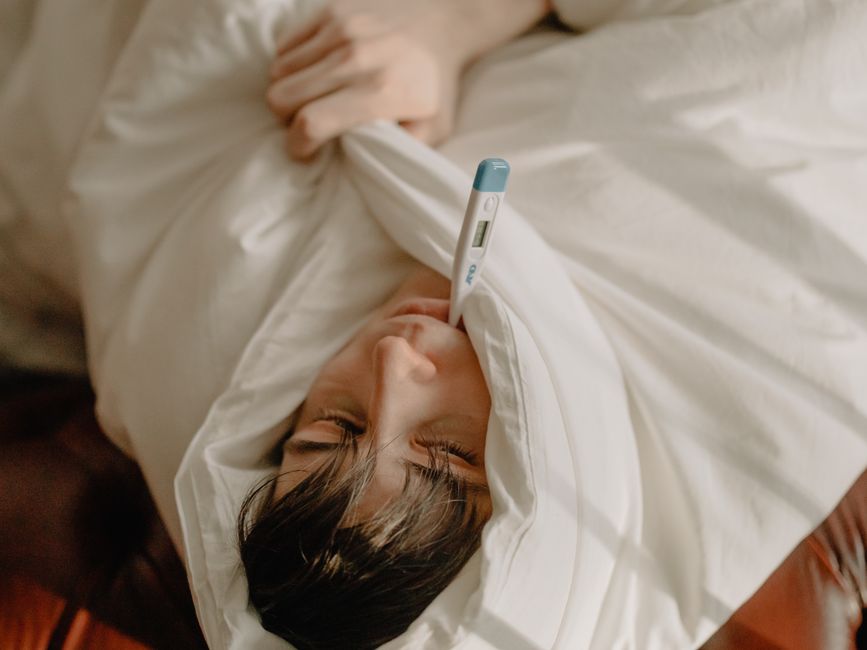 Sick man lying in bed, wrapped in blanket, holding a thermometer in mouth.