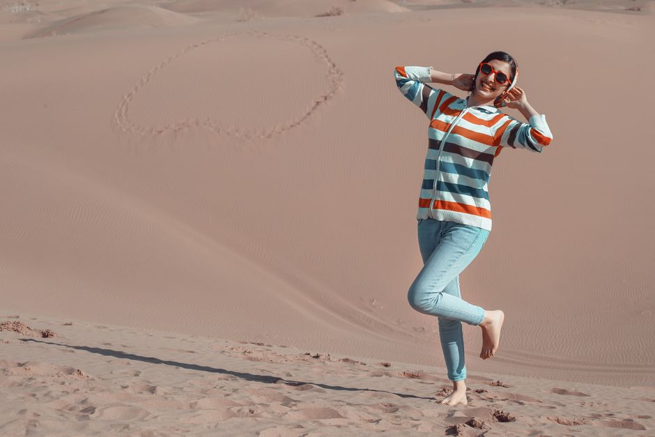 Cheerful young ethnic barefoot female traveler in casual wear and sunglasses touching hair while standing on one leg on sand with footprints during summer trip