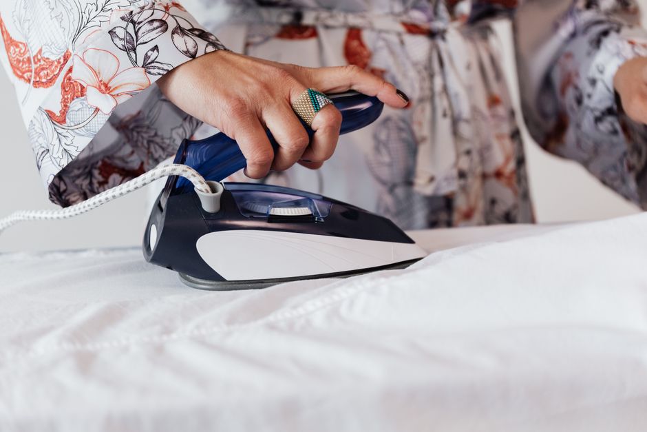 Crop anonymous housewife wearing elegant silk robe and stylish ring ironing white clothes with modern iron