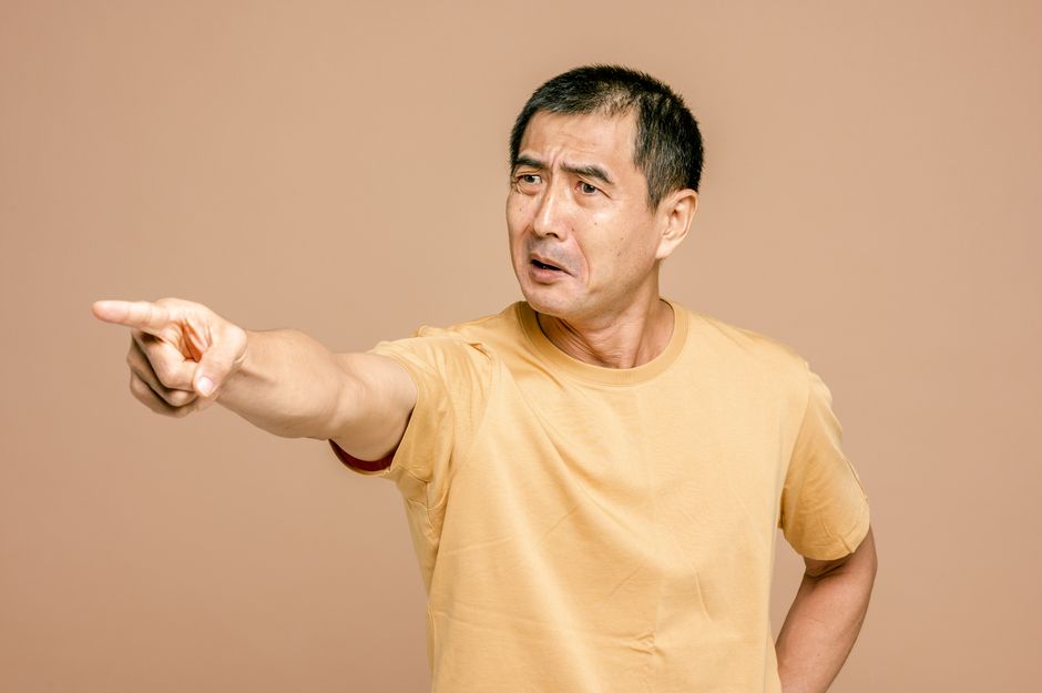 Senior Asian man expressing anger and pointing on a brown background.