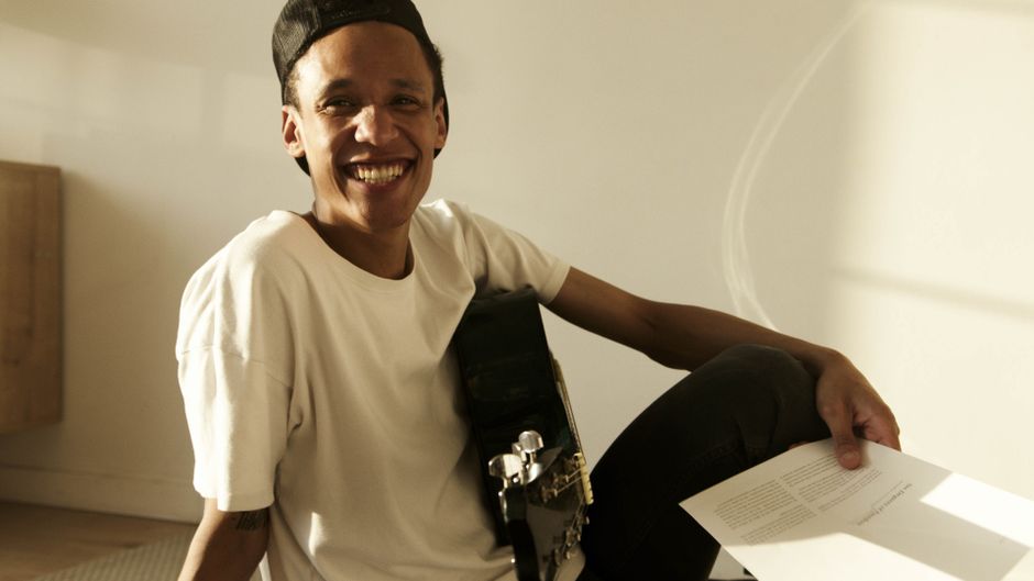 Young musician smiling while holding guitar and sheet music, exuding a relaxed lifestyle indoors.