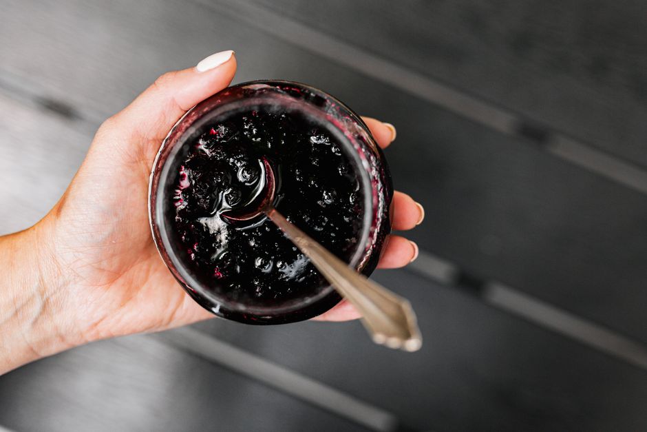 A hand holds a jar of homemade blackberry jam with a spoon, highlighting the rich and sweet texture.