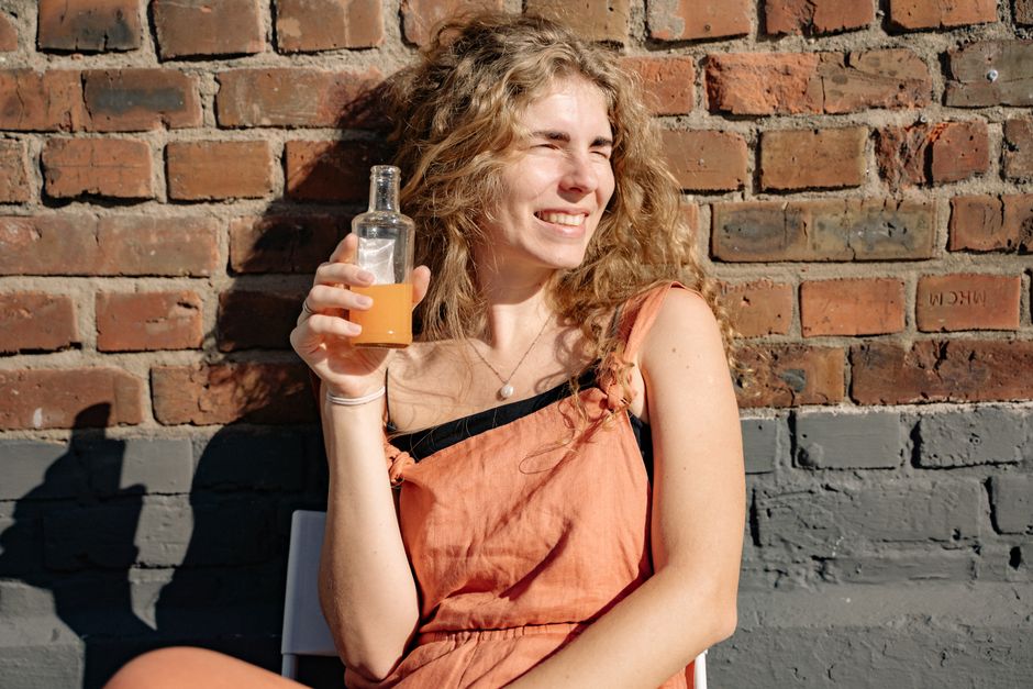 Smiling woman holding a drink, sitting against a brick wall in the sun.