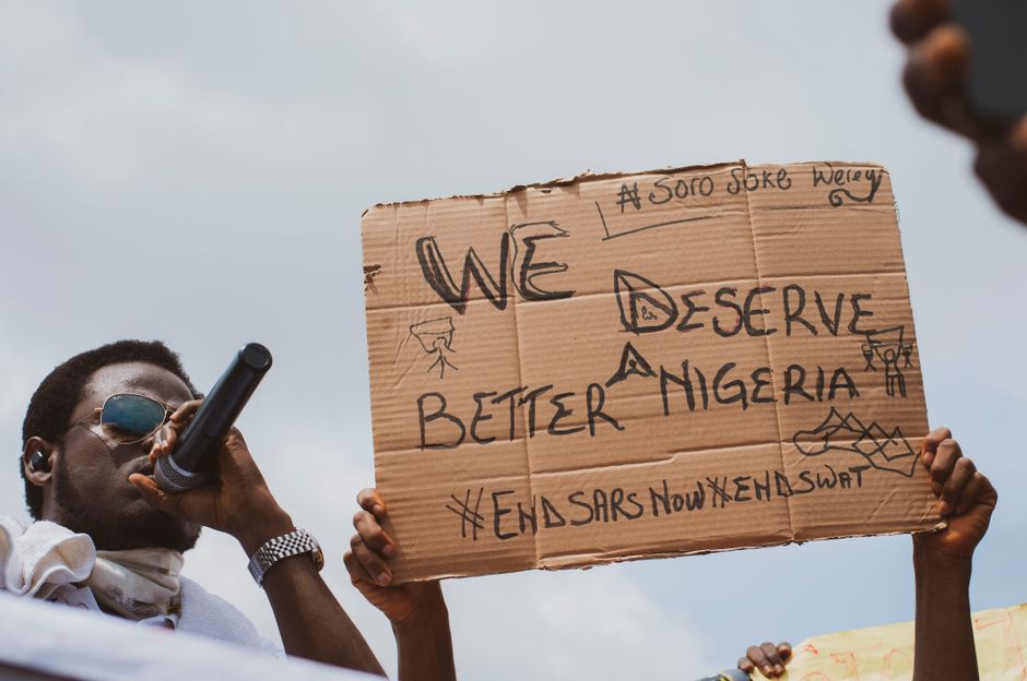 A Nigerian protester speaks into a microphone while holding a cardboard sign demanding a better future.