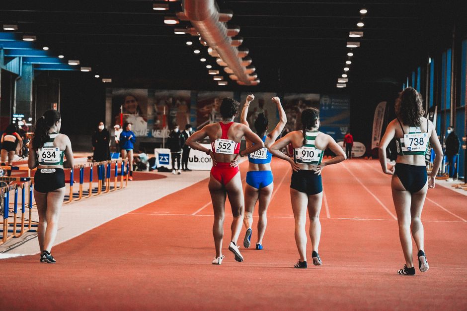 Group of female athletes at the starting line of an indoor track race, wearing numbered sportswear.
