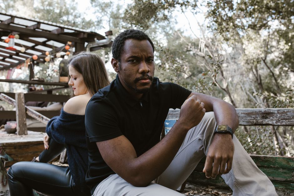 A thoughtful man sits outdoors, with a woman sitting distantly behind him.