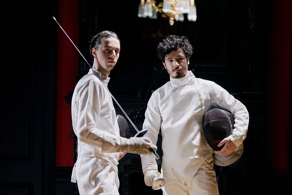 Two fencers in white gear posed confidently holding weapons inside a dim room.