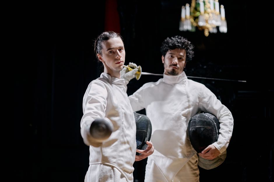 Two male fencers in protective gear strike a dramatic pose indoors.