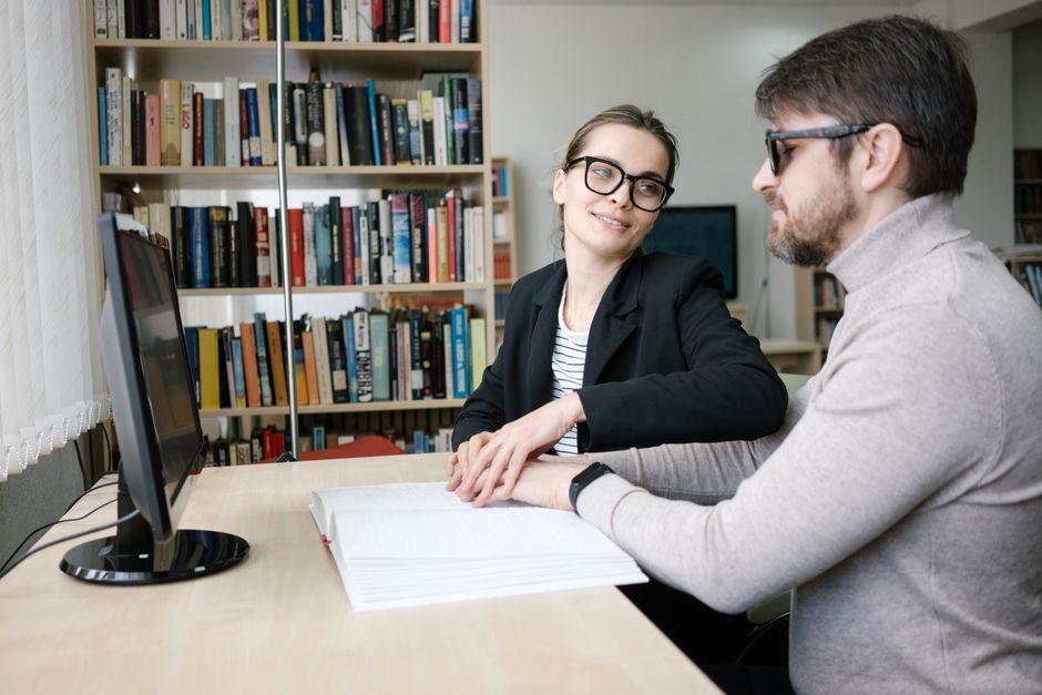 A woman assists a visually impaired man with reading Braille in a library setting.