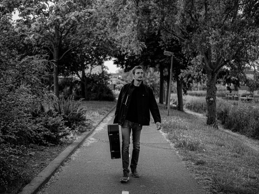 Black and white photo of a musician walking through a park with a guitar case.