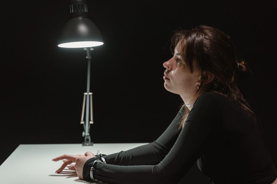 Side view of a woman sitting in an interrogation room under a lamp, handcuffed at desk.