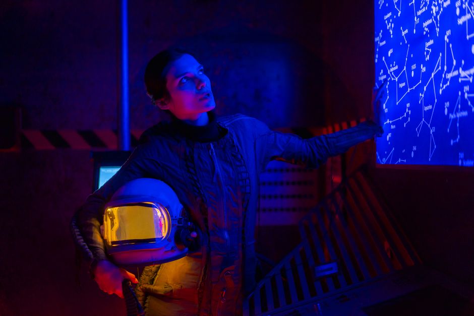 A spacewoman in a futuristic setting examines a star map, holding a space helmet under neon lighting.