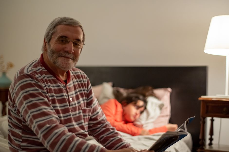 Senior man reading in a cozy bedroom while a young person rests nearby, capturing a warm family moment.