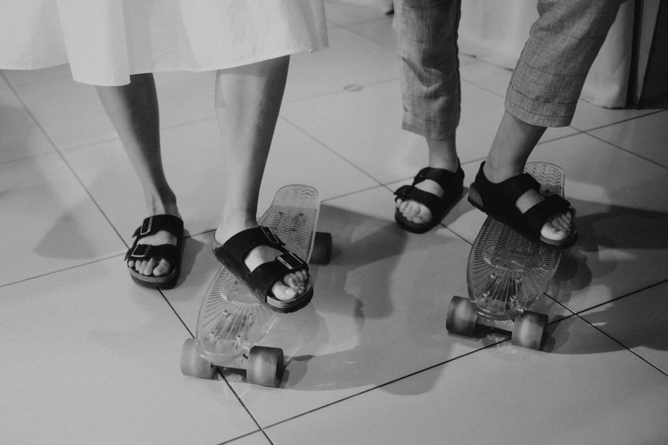 Monochrome image of two people wearing sandals standing on skateboards indoors.