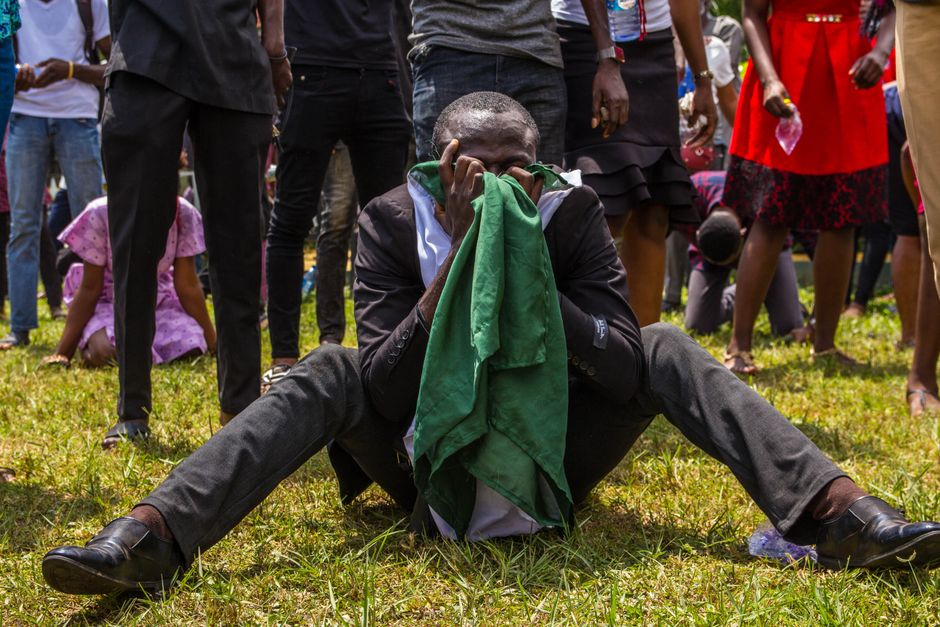 A man sitting on grass in emotional distress, obscured face, surrounded by a crowd outdoors.