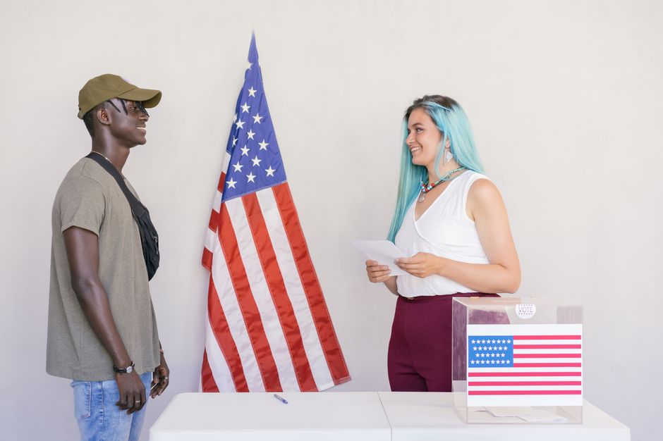 Two diverse young adults engaging in a voting process next to an American flag.