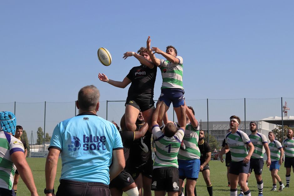 Athletes compete fiercely in an outdoor rugby match with players in green and black uniforms reaching for the ball.