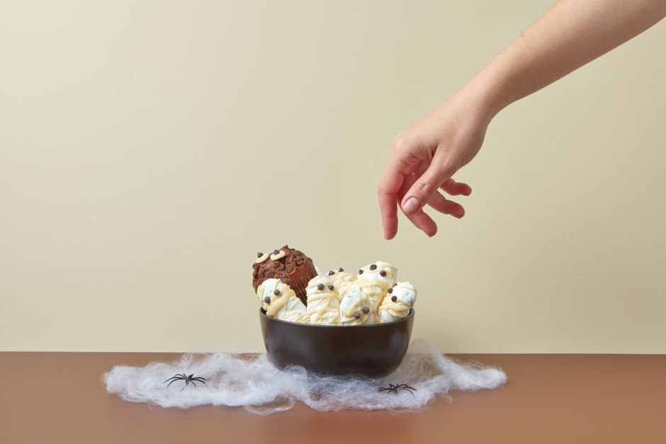 A hand reaching for a Halloween-themed dessert with mummy marshmallows in a bowl, set against a yellow background.
